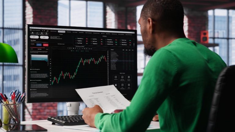 Man in green sweater sitting in front of a computer engaging in Stock trading — Forex, CFD, Derivatives.