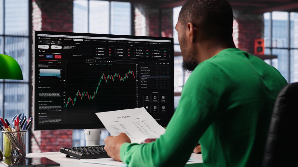 Man in green sweater sitting in front of a computer engaging in Stock trading — Forex, CFD, Derivatives.